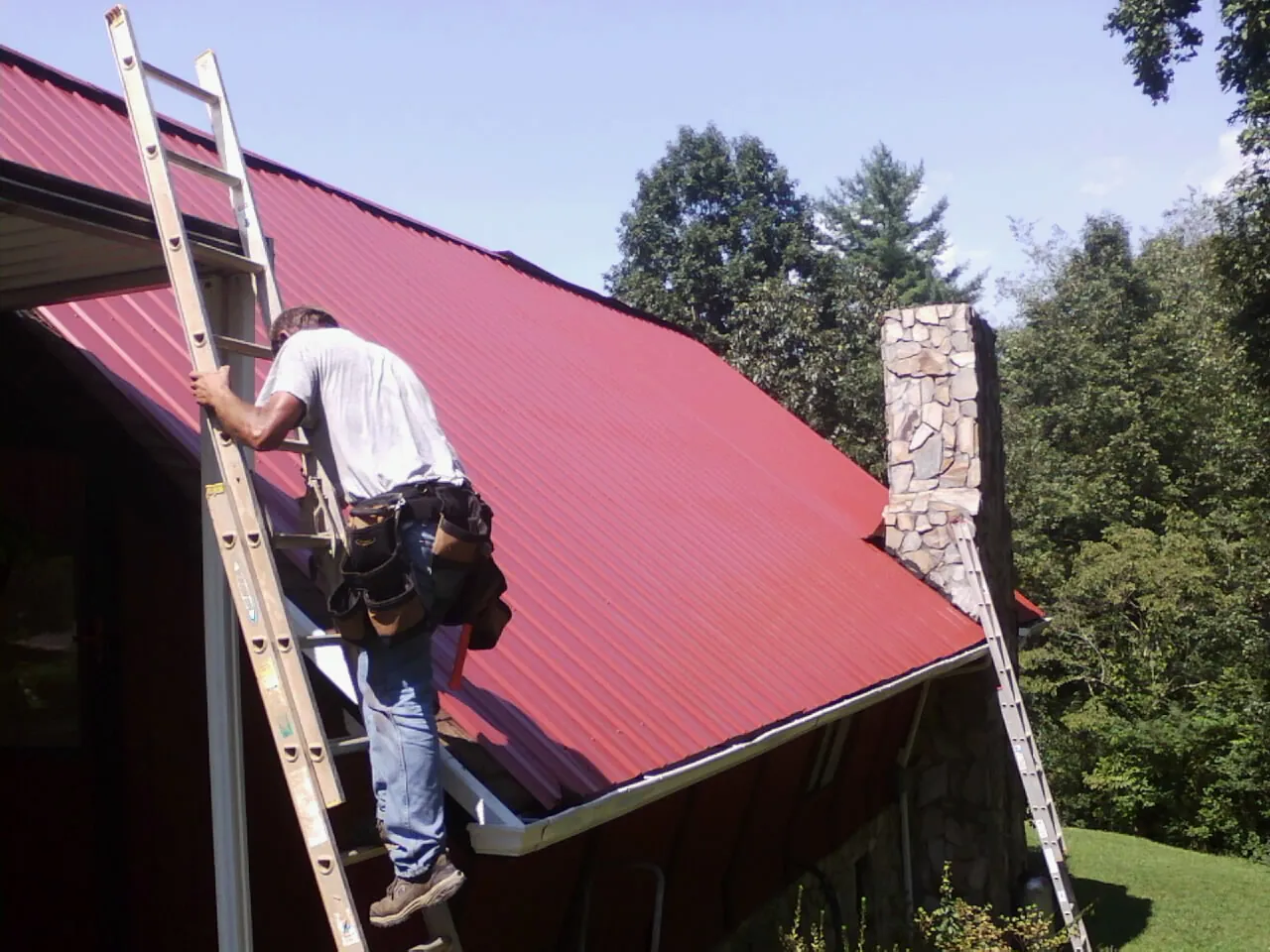 Roofer on a ladder beside a red metal roof and stone chimney — Soffit & Fascia Repair by Roofing Brothers in Festus